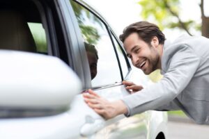 A man in a suit smiles as he closely examines the side of a white car outdoors on a sunny day, reviewing keywords.