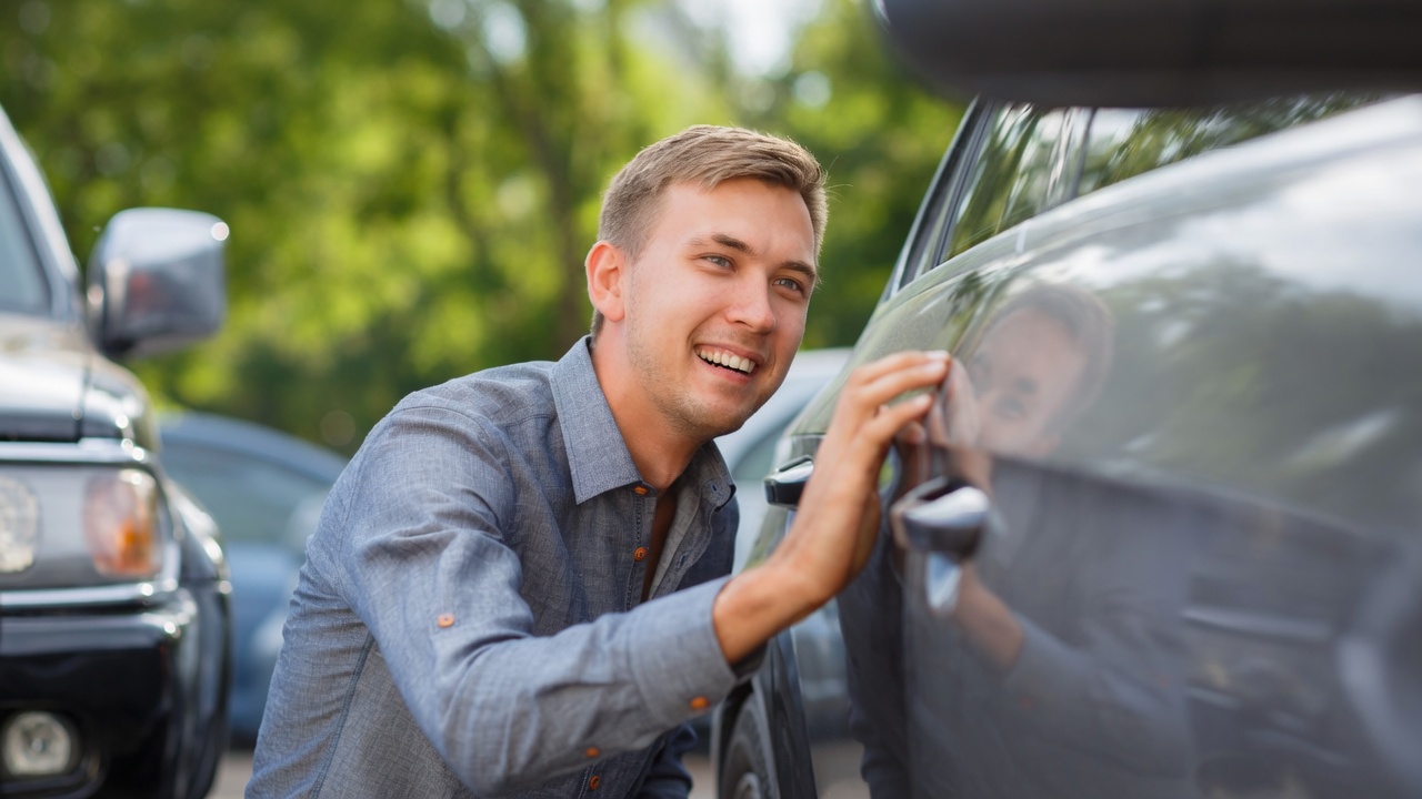 A man crouches by a car in an outdoor parking lot, smiling as he closely examines the side.