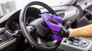 A gloved hand wipes a car steering wheel with a purple microfiber cloth.